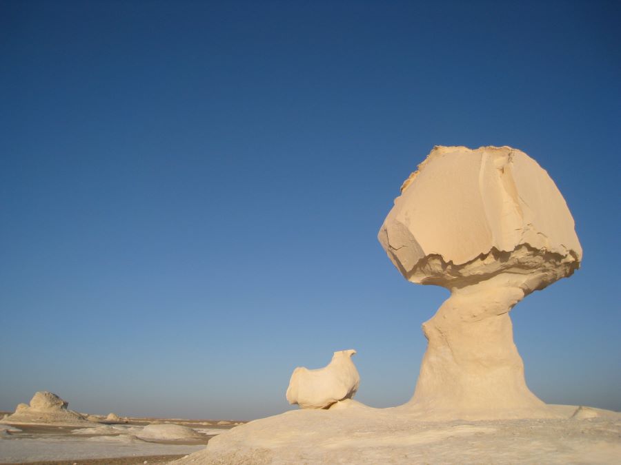 image of the white desert in egypt with two chalk formations pictured off-center in front of a clear blue sky, one chalk formation due to erosion looks like a chicken and the other looks like a bomb exploding, and these are commonly called the chicken and the bomb,